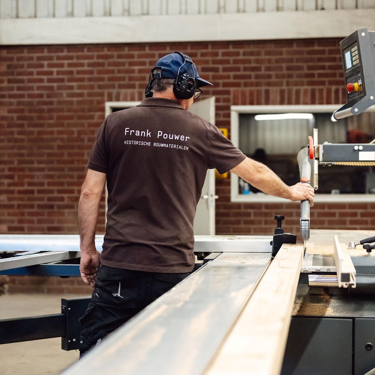 Medewerker zaagt een houten balk op maat in een houtwerkplaats, met gehoorbescherming en professionele zaagmachine.