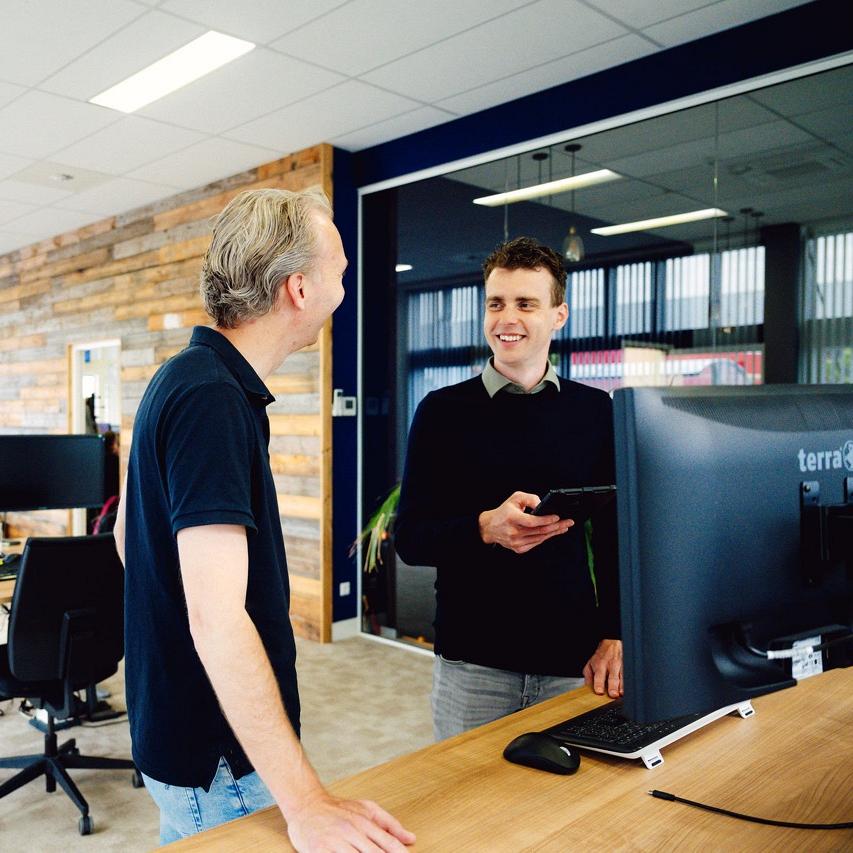 ChatGPT zei:  Twee mannen praten glimlachend bij een werkplek met dubbele monitoren in een modern kantoor met houten accentmuur, glazen vergaderruimte en veel daglicht.