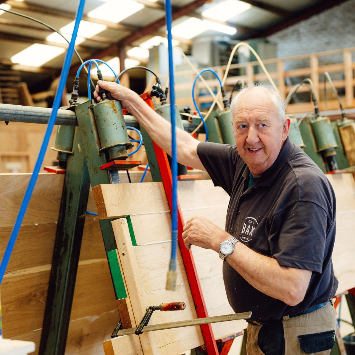 Vrouw met gehoorbescherming houdt houten latten vast in een houtwerkplaats en glimlacht tijdens haar werk.