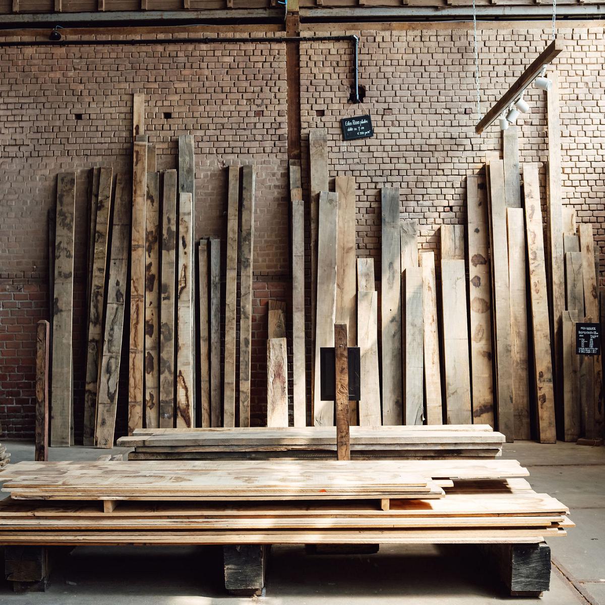 Houten planken en balken gestapeld in een werkplaats met een bakstenen muur en betonnen vloer.