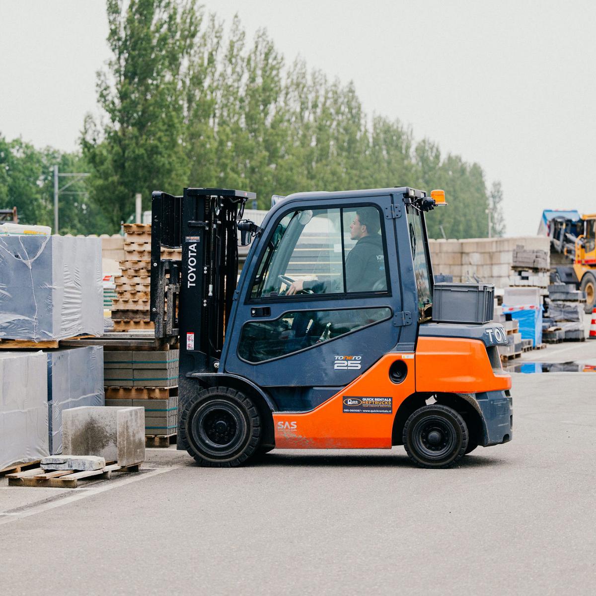 Oranje heftruck verplaatst een pallet met bouwmaterialen op een buitenterrein, omringd door gestapelde stenen en andere machines op de achtergrond.