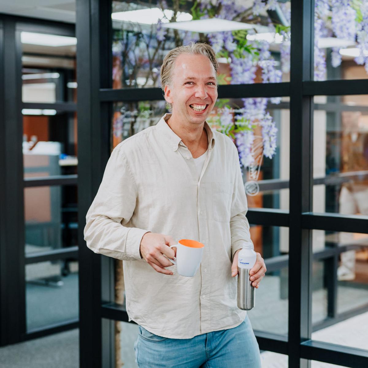 Lachende man in lichte outfit staat met een mok en drinkbeker in de hand in een moderne kantoorruimte met glazen wanden.