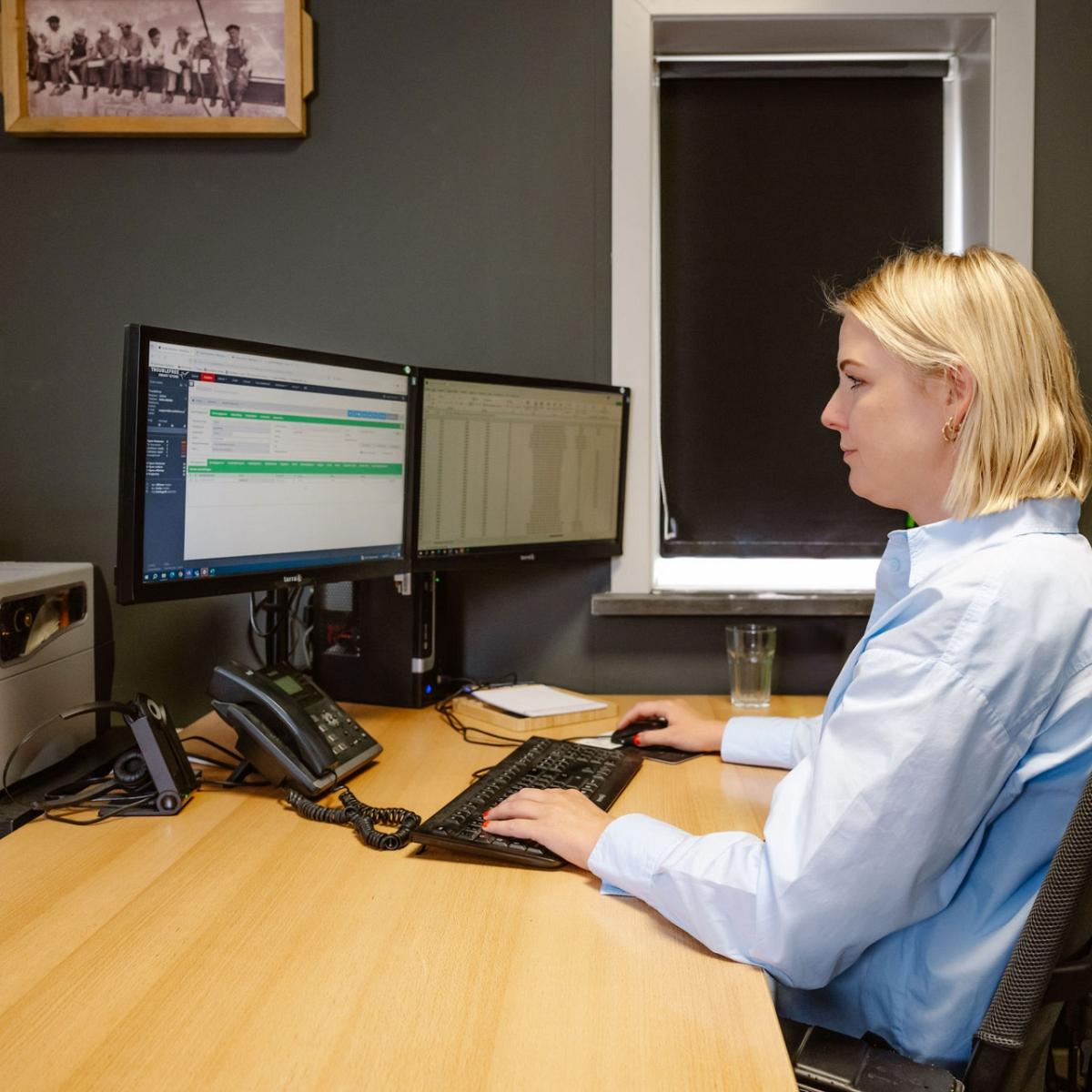 Vrouw in een lichtblauwe blouse werkt op een computer met twee schermen in een kantooromgeving, met een telefoon, printer en glas water op het bureau.