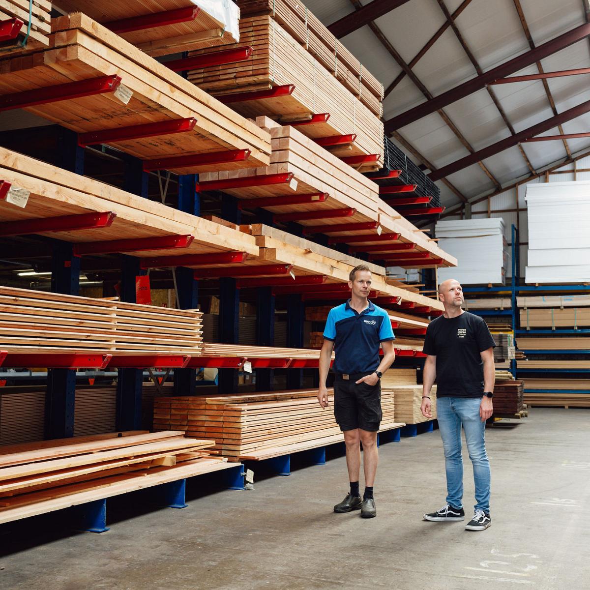 Twee medewerkers in een magazijn met hoge stellingen vol hout en plaatmateriaal.