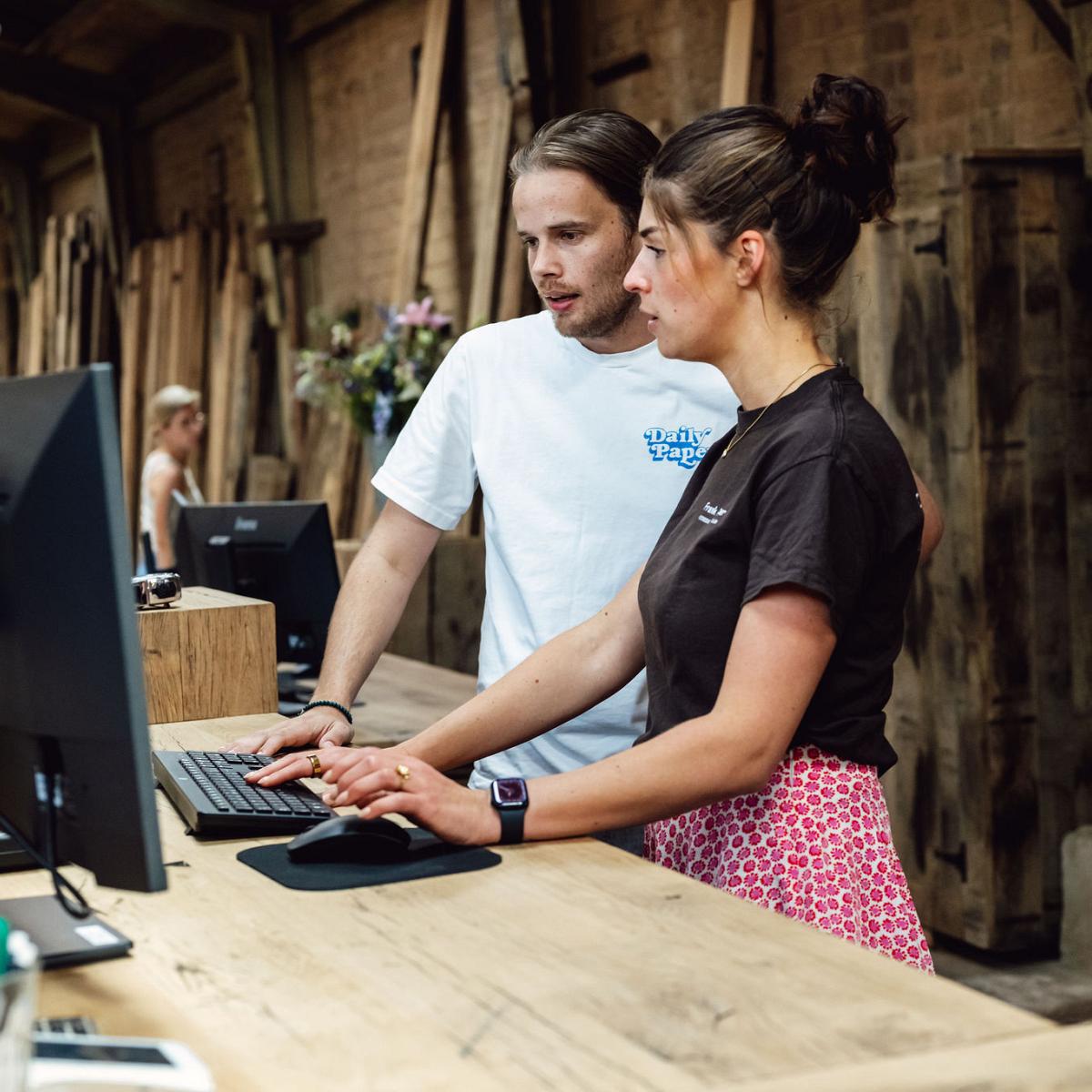 Consultant en  collega overleggen achter een houten balie met computers in een werkplaats met houten planken op de achtergrond.