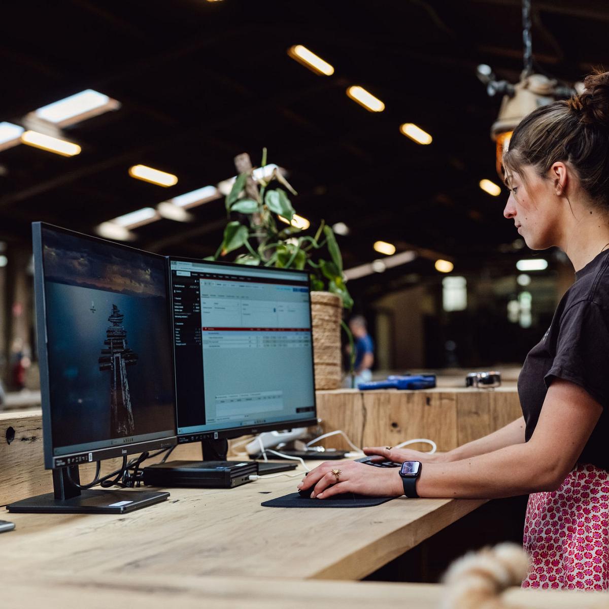 Vrouw werkt staand achter een houten balie met twee beeldschermen in een ruime, ambachtelijke werkplaats.