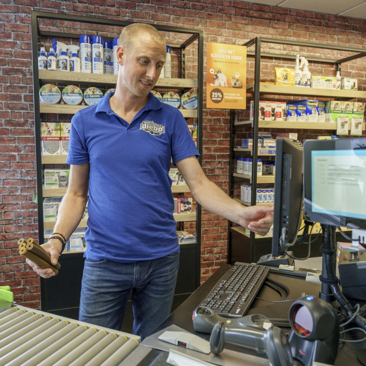 Een man achter een winkeltoonbank met verschillende producten op de planken en een computerkassa.