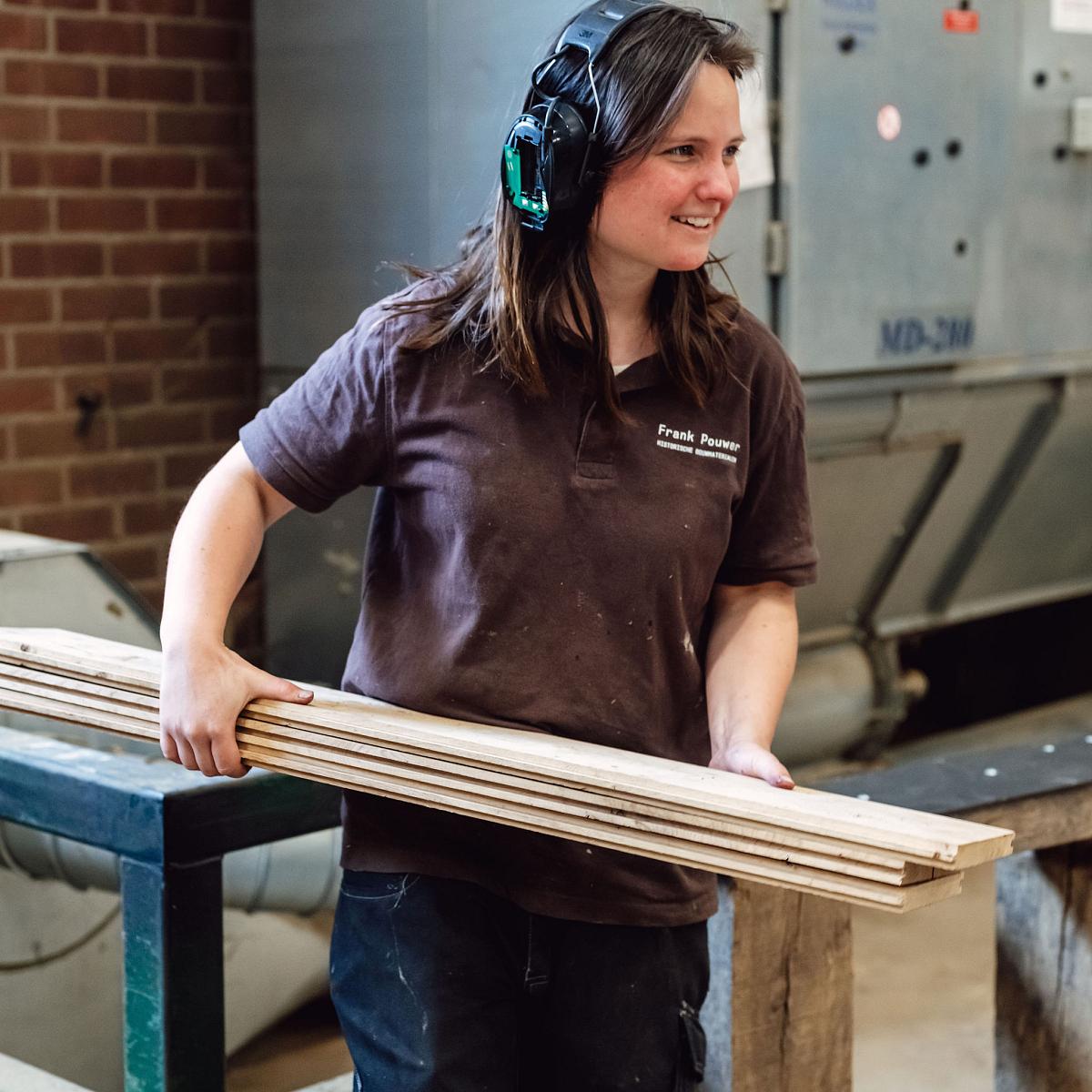 Vrouw met gehoorbescherming houdt houten latten vast in een houtwerkplaats en glimlacht tijdens haar werk.