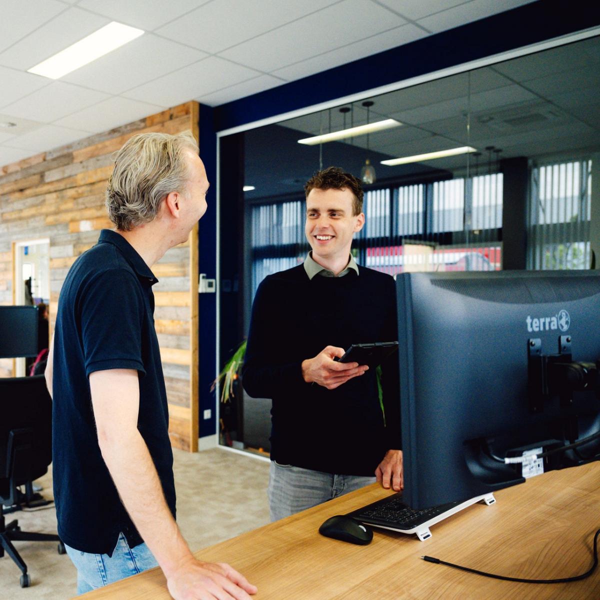 Twee personen praten met elkaar in een kantooromgeving met bureaus, computers en monitors.