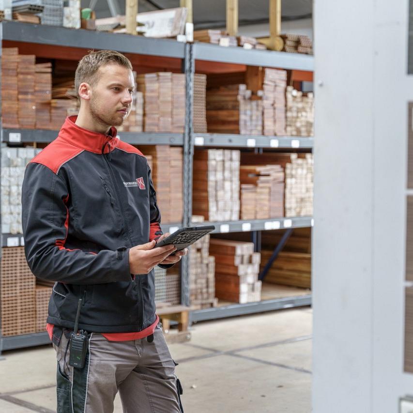 Man staat met tablet in een houtloods met stellingen met hout op de achtergrond.
