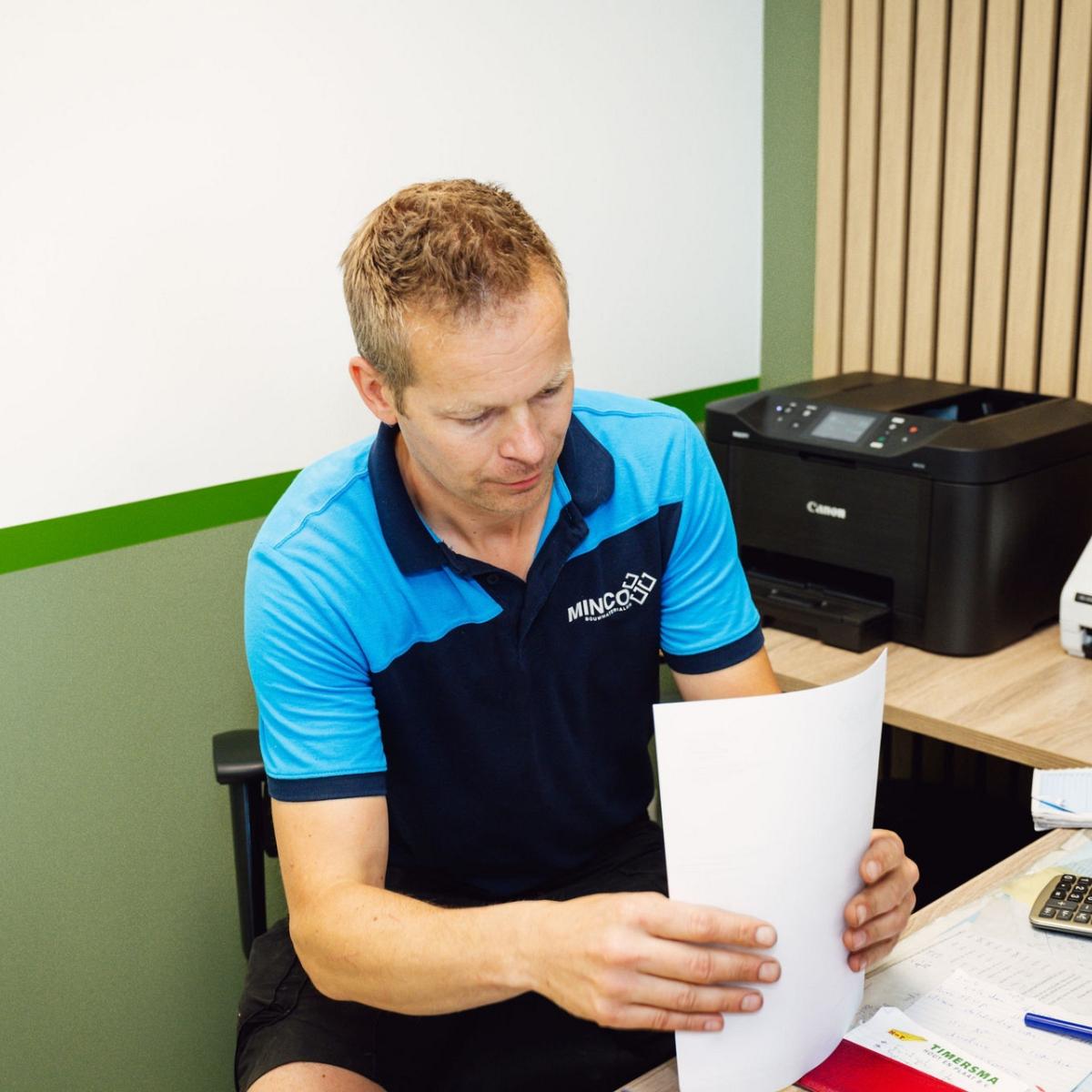 Man met papieren in handen aan een bureau met daarop printer, rekenmachine en telapparaat.
