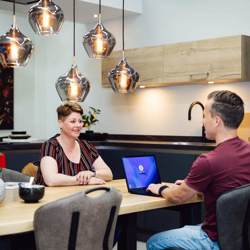 Een man zit achter zijn laptop terwijl hij in gesprek is met een vrouw aan tafel.