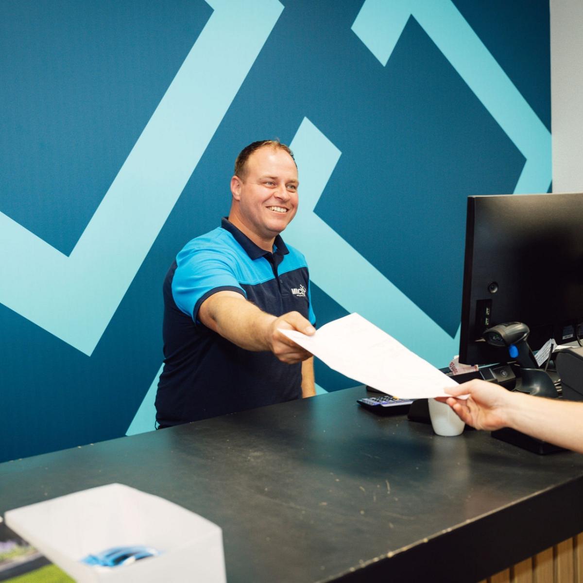 Een hand geeft een formulier aan een man die achter een balie staat.