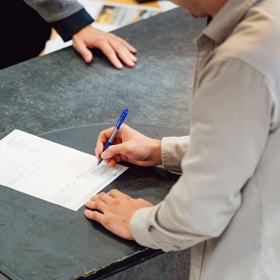 Persoon ondertekent een formulier aan een balie met op de balie een hand van ander persoon.