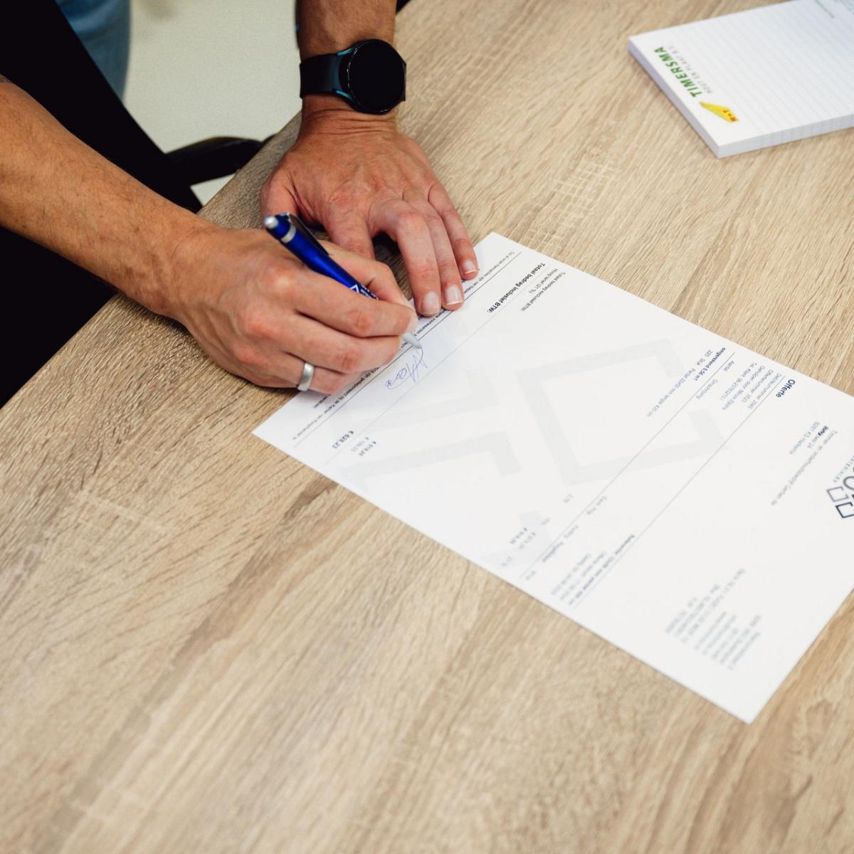 Twee handen ondertekenen een formulier op een houten tafel.