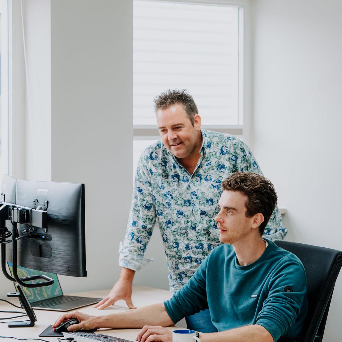 Twee collega’s werken samen achter een bureau; de een zit achter de computer terwijl de ander naast hem staat en meekijkt op het scherm.