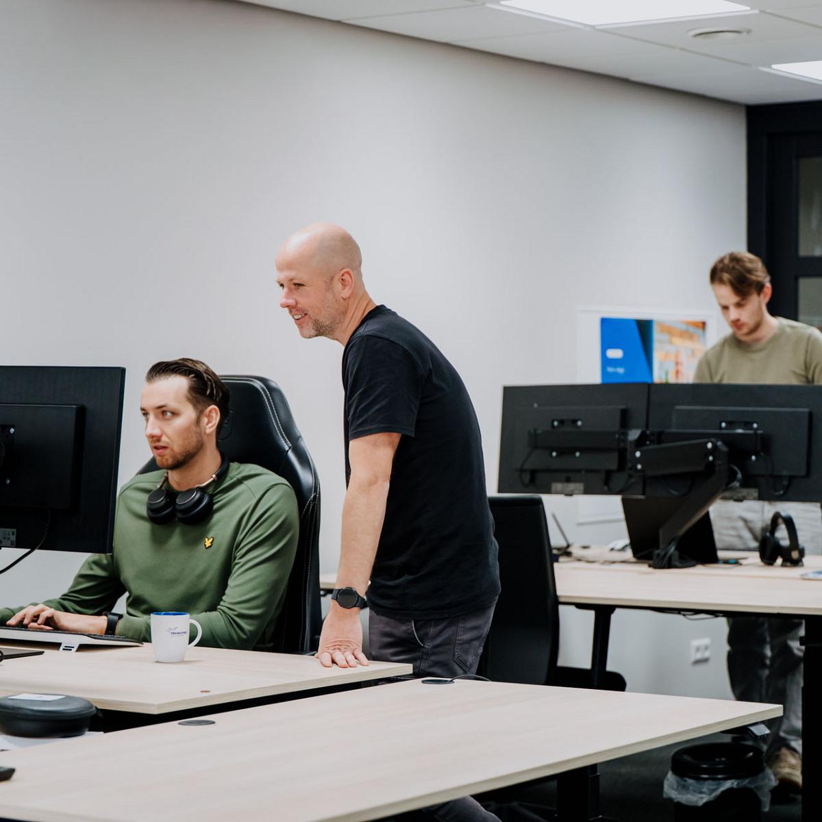 Drie collega’s aan het werk op kantoor; twee overleggen achter een computerscherm terwijl een derde op de achtergrond bij zijn bureau werkt.