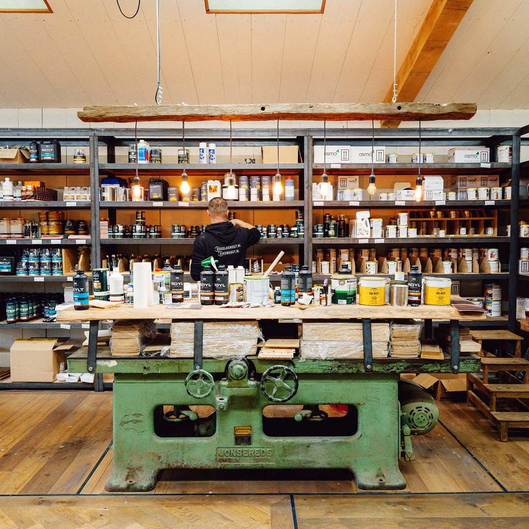 Oude werkbank met verschillende bouwmaterialen hierop. Op de achtergrond stellingen waar bouwmaterialen in staan en een man die iets pakt uit de stelling.