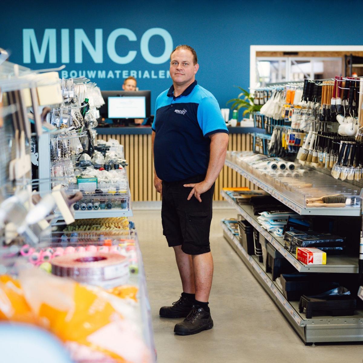 Een man in een gangpad van een winkel vol met bouwmaterialen met een blauwe want met het minco logo op de achtergrond.