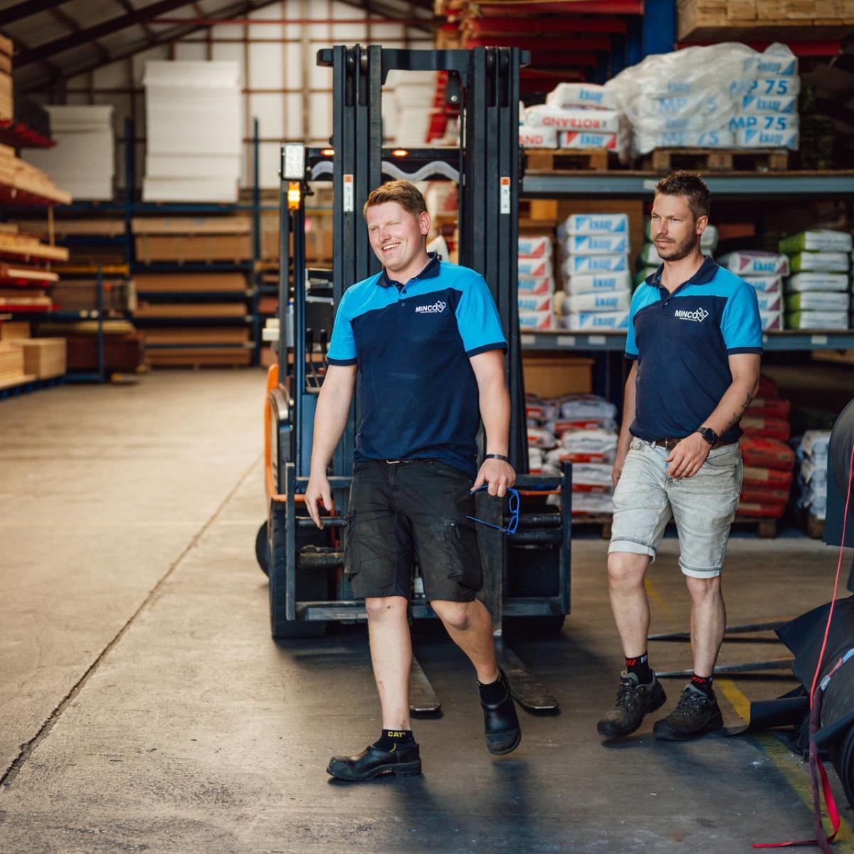 lachende mannen in een magazijn vol houten balken met een heftruck op de achtergrond.