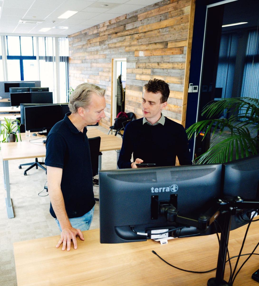 Twee mannen overleggen bij een werkplek met dubbele monitoren in een modern kantoor met houten wand, planten en grote ramen met veel daglicht.