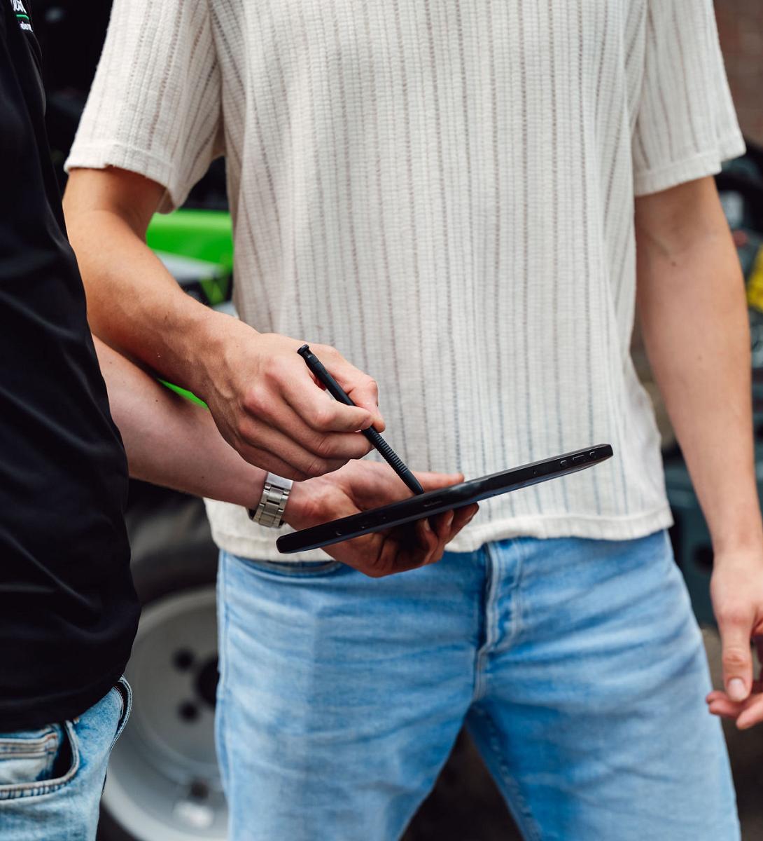 Twee mannen overleggen buiten terwijl één van hen met een stylus gegevens invoert op een tablet, met landbouwvoertuigen op de achtergrond.