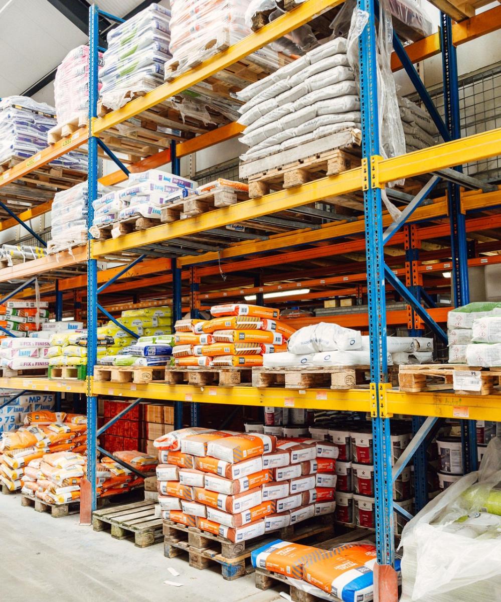 Opslagruimte met hoge stellingen gevuld met bouwmaterialen zoals cementzakken, isolatiemateriaal en verpakte producten, georganiseerd op pallets.