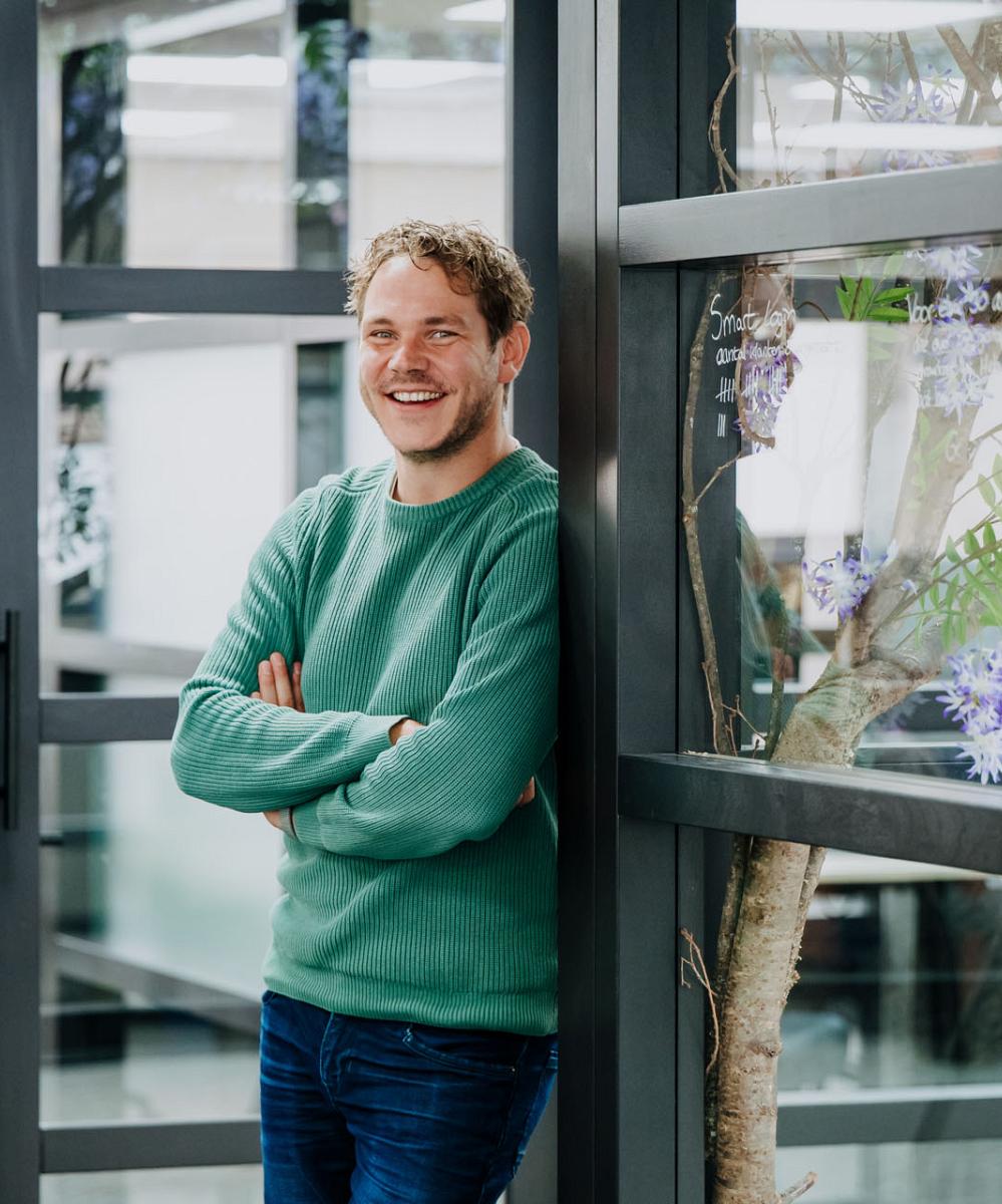 Lachende man in groene trui leunt ontspannen tegen een glazen wand in een moderne kantoorruimte met planten en bureaus op de achtergrond.