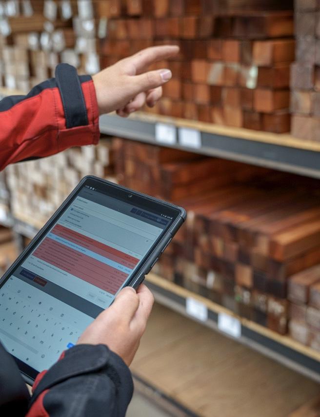 Handen die een tablet vasthouden met daarop Smart Trade software en de andere hand wijst naar een stelling met hout blokken. 
