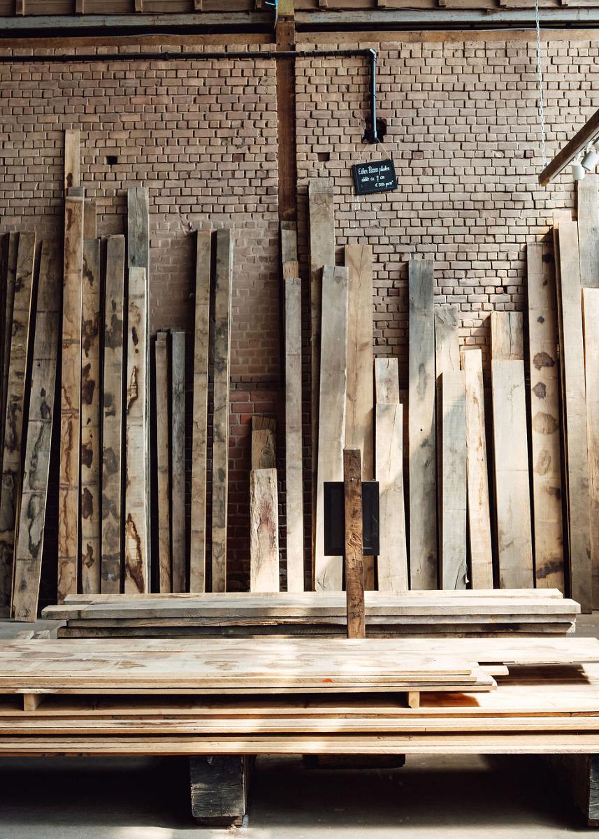 Houten planken en balken gestapeld in een werkplaats met een bakstenen muur en betonnen vloer.