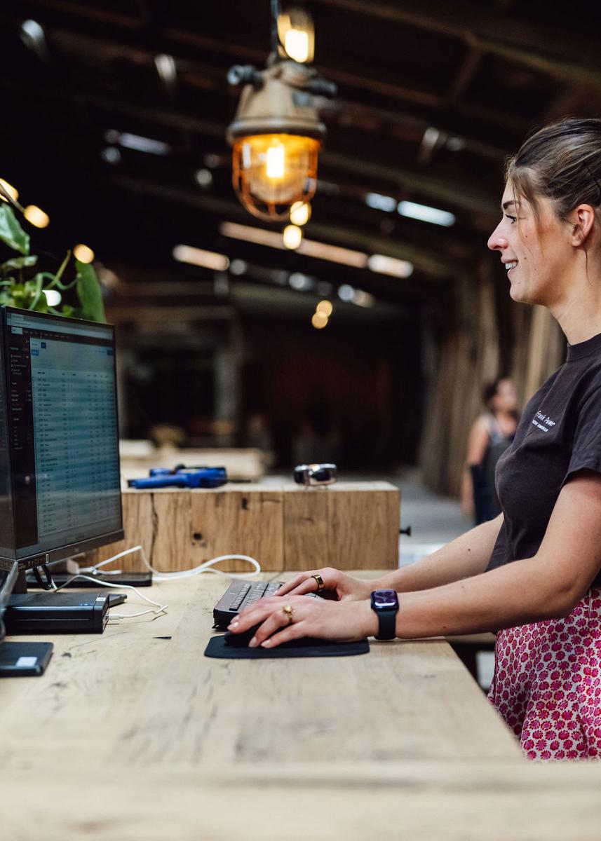 Vrouw werkt staand achter een houten balie met twee beeldschermen in een sfeervolle houtloods met warme verlichting en houten details.