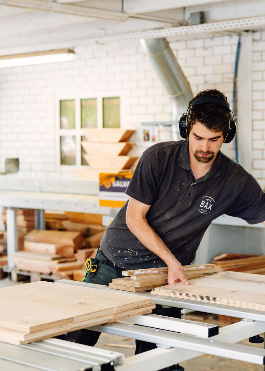 Man in werkshirt en gehoorbescherming zaagt houten platen op maat in een timmerwerkplaats, omringd door houtmateriaal en gereedschap.