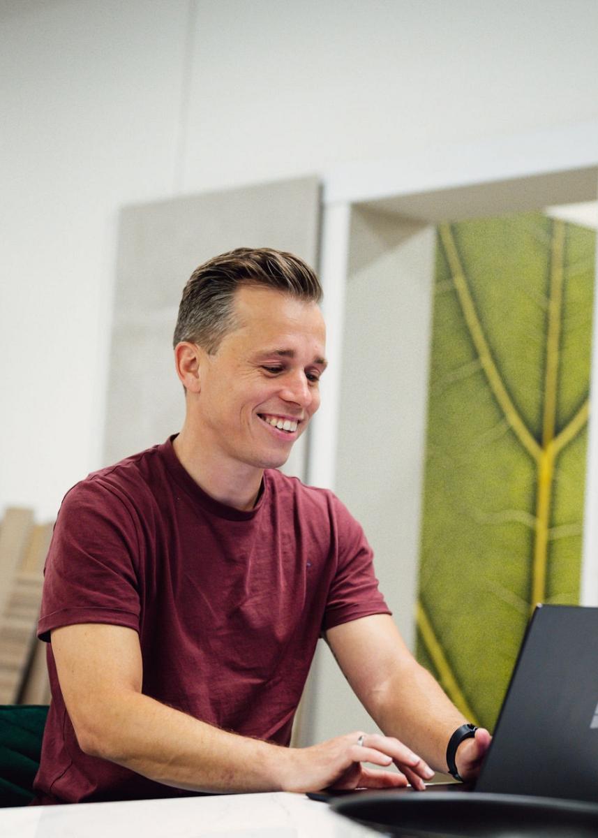 Man in een rood T-shirt werkt glimlachend op een laptop in een interieurshowroom, omringd door staaltjes van hout, tegels en decoratieve panelen.
