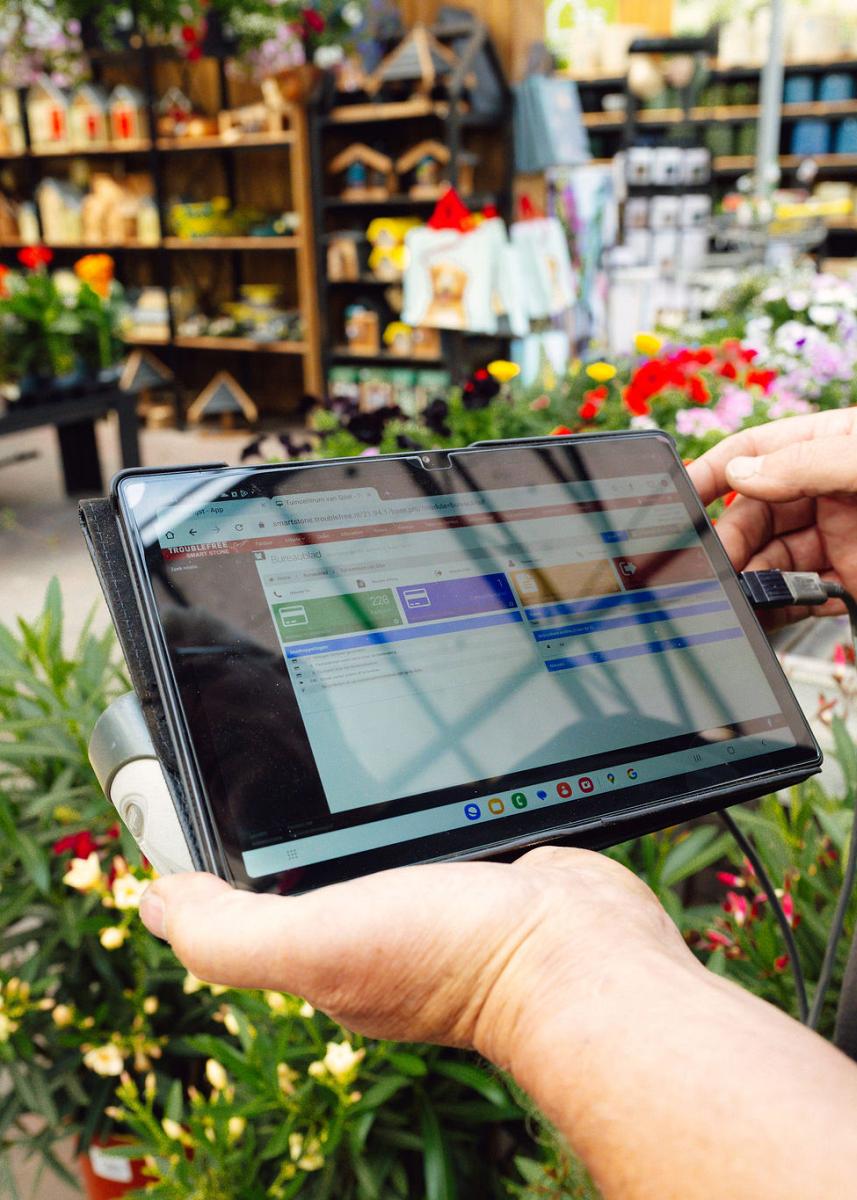 Een persoon houdt tablet in handen met informatie in een bloemenwinkel, met kleurrijke planten op de achtergrond.