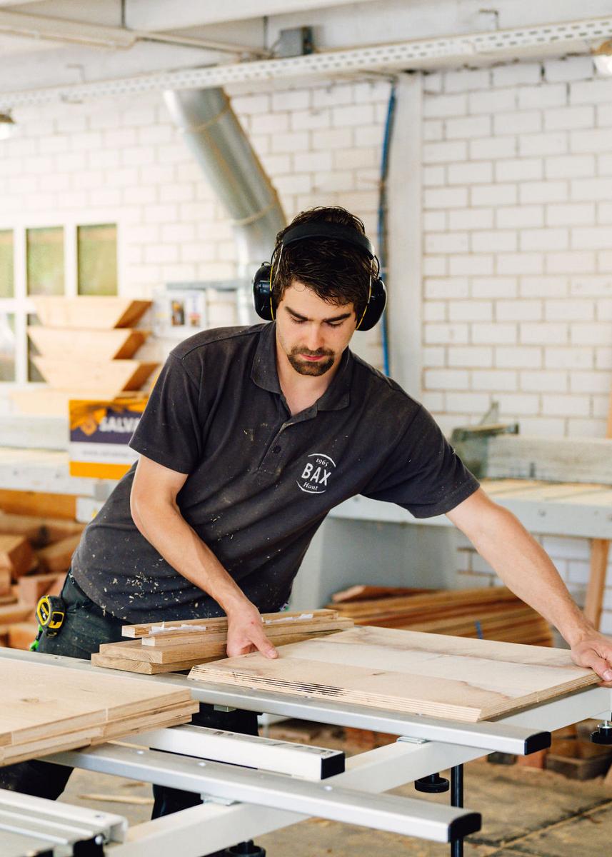 Man in werkshirt en gehoorbescherming zaagt houten platen op maat in een timmerwerkplaats, omringd door houtmateriaal en gereedschap.
