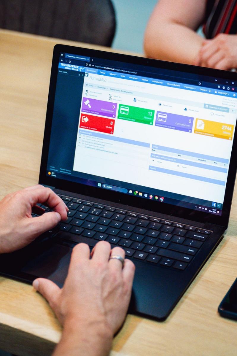 Laptop op tafel waar een man op aan het werk is met daarnaast een telefoon en aan de andere kant van de tafel zit een vrouw met haar handen over elkaar.