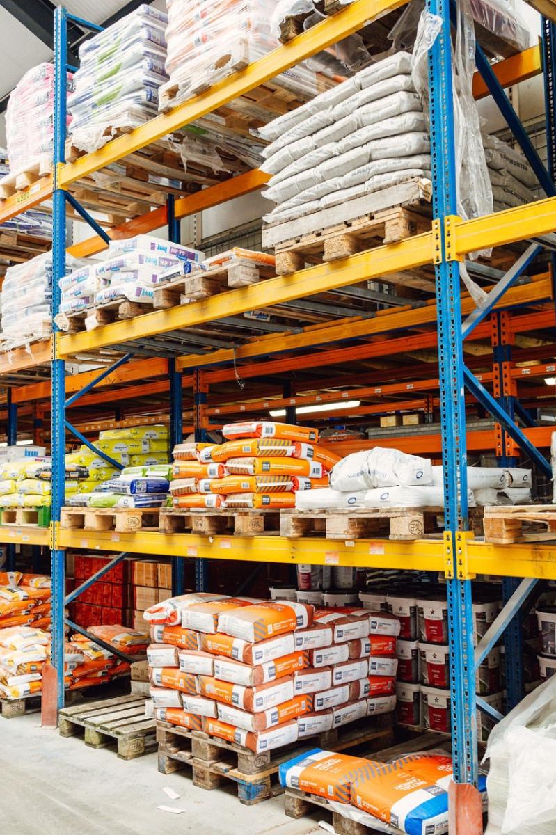 Opslagruimte met hoge stellingen gevuld met bouwmaterialen zoals cementzakken, isolatiemateriaal en verpakte producten, georganiseerd op pallets.