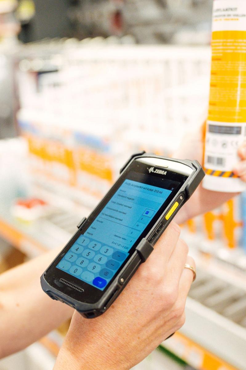Persoon scant een koker afdichtmiddel met een mobiele scanner in een winkel, met schappen vol vergelijkbare producten op de achtergrond.