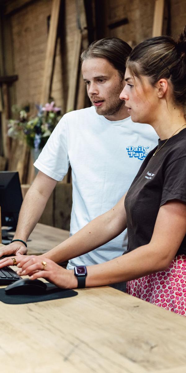 Consultant en  collega overleggen achter een houten balie met computers in een werkplaats met houten planken op de achtergrond.