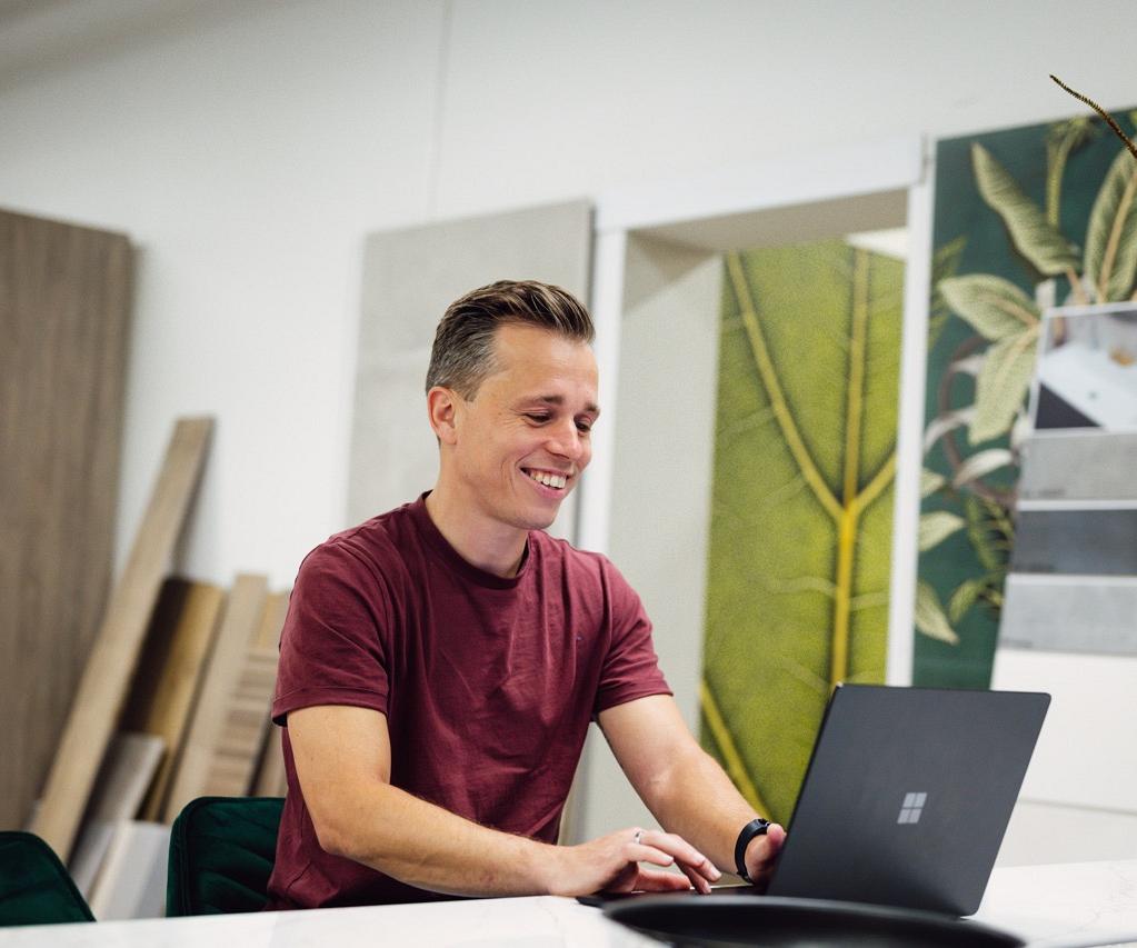 Man zit achter bureau met laptop voor zich waarop hij kijkt al glimlachend in een winkelomgeving.
