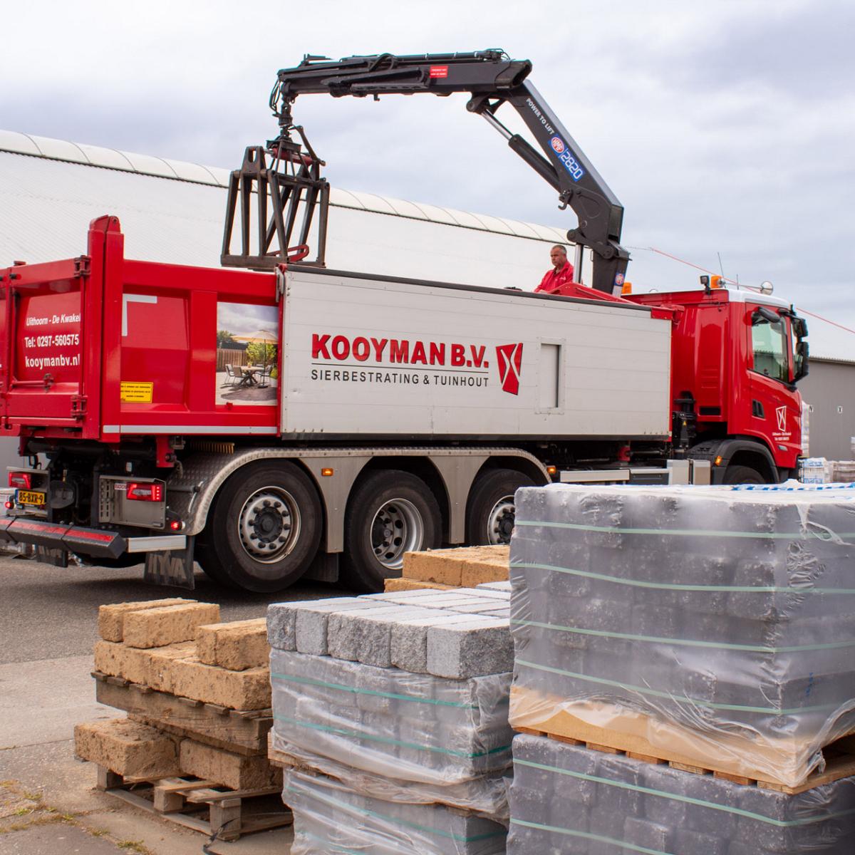 Vrachtwagen van Kooyman B.V. met kraan tussen pallets met bestratingsmaterialen