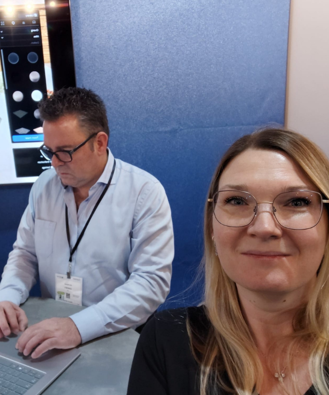 Vrouw met bril maakt een selfie op een beursstand, terwijl een man met badge achter haar op een laptop werkt bij een presentatie met ontwerptool op een scherm.