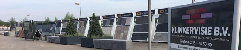 Zwart bord met 'Klinkervisie B.V.' erop. Op de achtergrond staan displays met verschillende tegels hierop. Hiervoor zijn plantenbakken te zien.