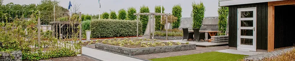 Showtuin van De Klinker Sierbestrating, met aan de linker kant een zwart tuinhuisje met overkapping, in het midden een ophoging met gras en daar omheen verschillende soorten stenen.