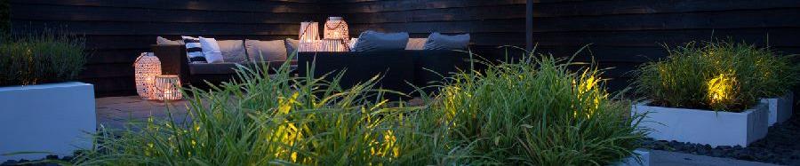 Twee witte plantenbakken op de voorgrond in een tuin met op de achtergrond een lounge set met daaromheen en hek met lichten.