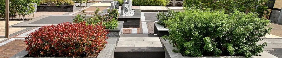 Twee grote vierkanten plantenbakken met in het midden een klein trappetje. Links heeft een rode struik en rechts groen. Op de achtergrond staan verschillende plantenbakken en tegels in display.