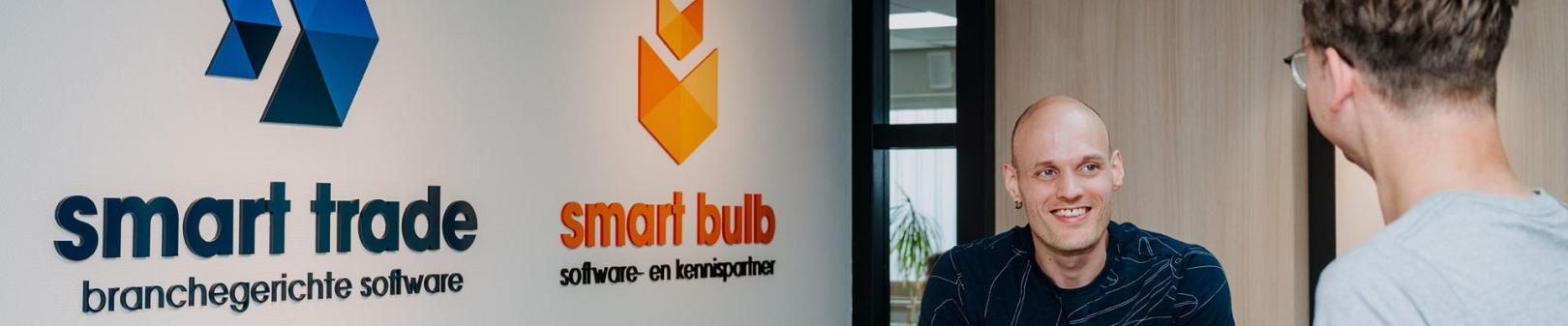 Medewerkers in gesprek aan een hoge tafel op kantoor, met de logo’s van SmartTrade en Smart Bulb zichtbaar op de muur op de achtergrond.