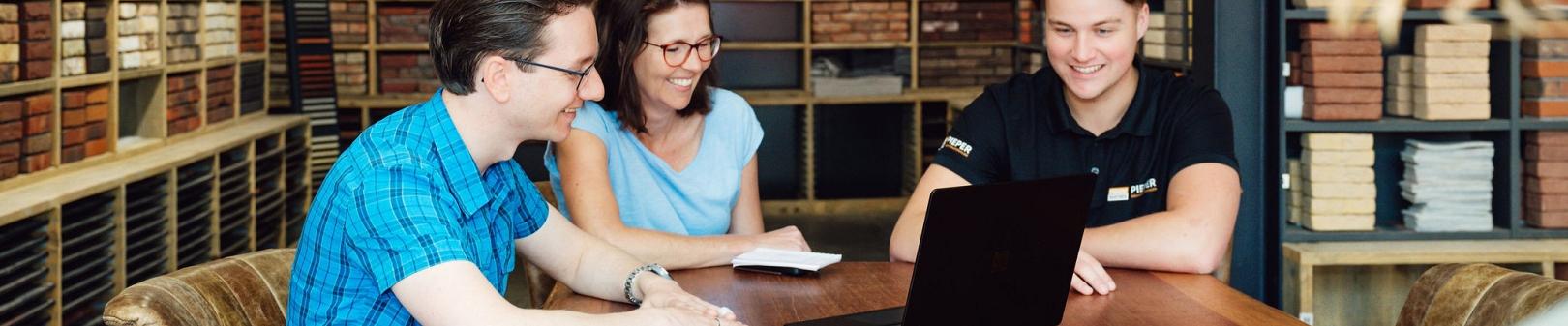 Drie professionals zitten aan een houten tafel in een showroom met baksteenmonsters en bekijken samen een laptop, omringd door wanden met diverse steensoorten en kleuren.