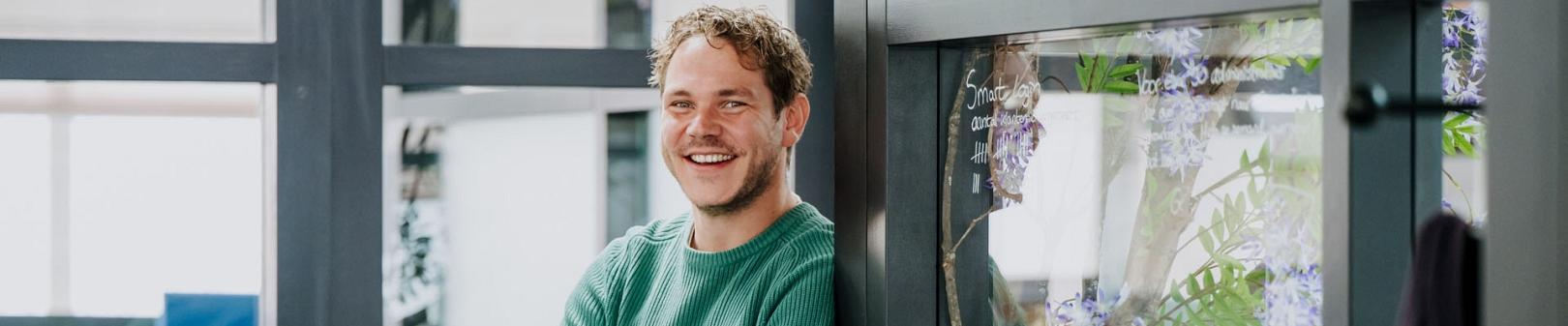 Medewerker van Smart Trade staat glimlachend met gekruiste armen in een moderne kantoorruimte met glazen wanden en een plant met paarse bloemen op de achtergrond.