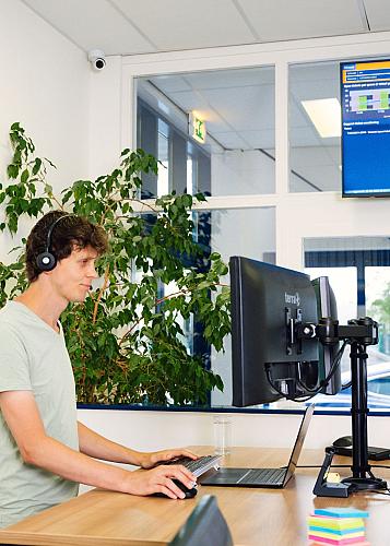 Man met koptelefoon die achter zijn bureau staat en aan het werk is op zijn computer met om de achtergrond een tv met statistieken en grote plant. 
